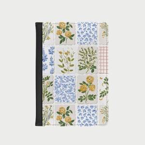 Op de afbeelding: Een paspoorthouder met een bloemenpatchworkontwerp. Het ontwerp bevat gele en blauwe bloemen, groen blad en een roze ruitpatroon. Het heeft een zwarte rug.