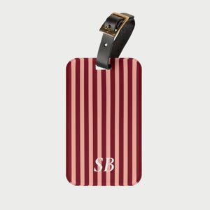 Op de afbeelding: Een bagagelabel met een verticaal gestreept patroon in rood- en roze tinten. Het label heeft een zwarte leren riem met een goudkleurige gesp. Witte initialen "SB" staan in het midden van het label.