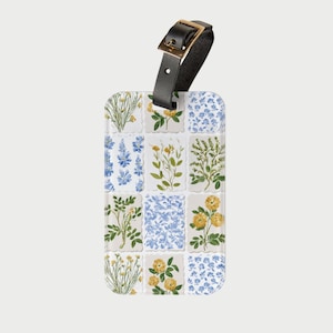 Op de afbeelding: Een rechthoekig bagagelabel met een zwarte leren riem en een gouden gesp. Het label heeft een bloemenpatroon met blauwe en gele bloemen op een witte achtergrond. Ontworpen voor reizen.