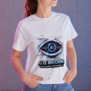 May include: White t-shirt featuring a graphic of an eye with the letters "AI" in the center. The text "AI IS WATCHING (AND JUDGING YOUR PROMPTS)" is below the eye graphic. The shirt is made of a soft material.