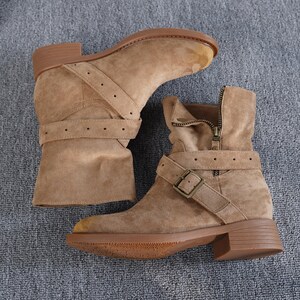 May include: Tan suede ankle boots with a low heel. The boots have a side zip, buckle straps, and a rounded toe. The soles and heels are brown. The boots are approximately 10 cm high.