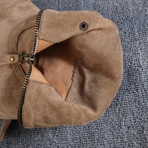 May include: Close-up of a tan suede boot with a zip and buckle detail. The boot's interior is visible, revealing a lighter tan lining. The zip is partially open, showcasing the boot's construction and materials.