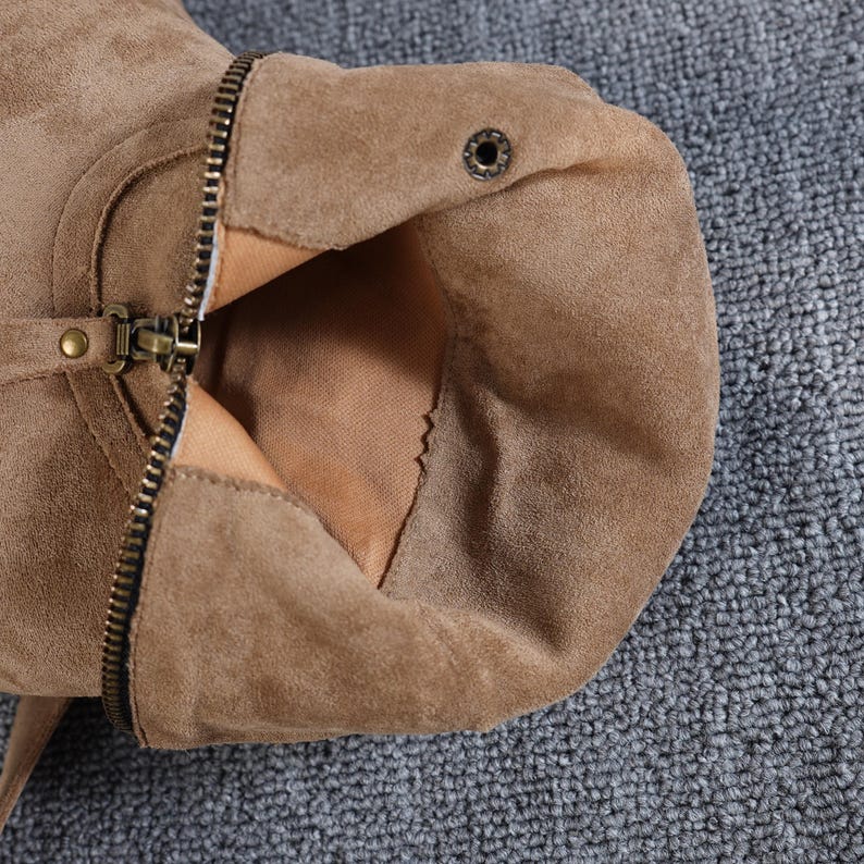 May include: Close-up of a tan suede boot with a zip and buckle detail. The boot's interior is visible, revealing a lighter tan lining. The zip is partially open, showcasing the boot's construction and materials.