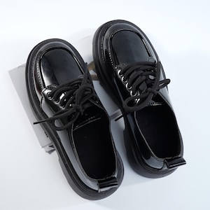 May include: Pair of glossy black platform shoes with laces. The shoes have a rounded toe and a thick sole. The laces are black and are threaded through silver eyelets. The shoes are sitting on a white surface.