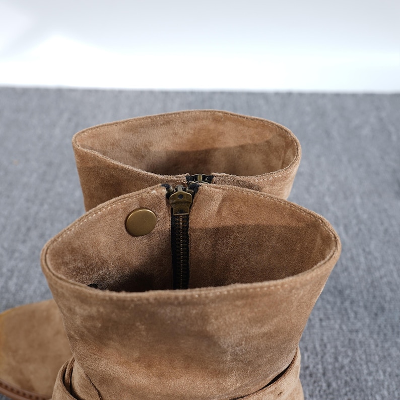 May include: Close-up of tan suede ankle boots. The boots feature a zip closure and a decorative button. The interior of the boots is visible, showing the soft suede material. The boots are a neutral colour, suitable for various outfits.