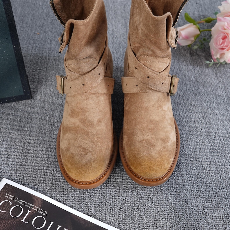 May include: Tan suede ankle boots with a round toe and low heel. The boots have a side zip, adjustable buckle straps, and a stitched sole. The boots are displayed on a grey surface.