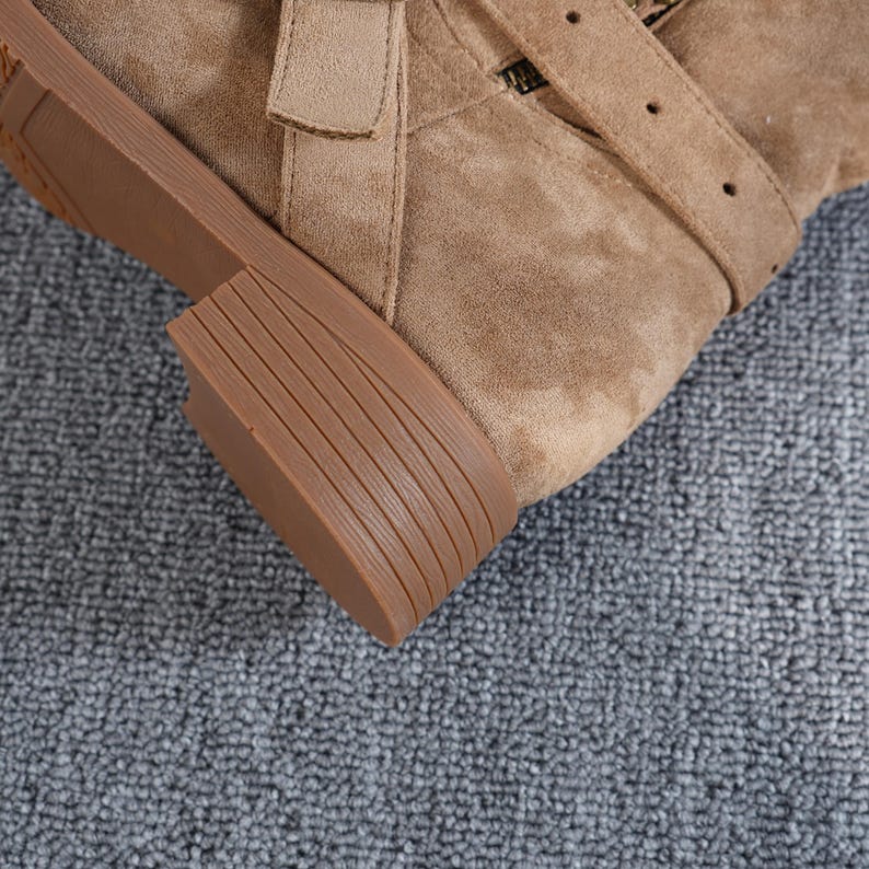 May include: Close-up of a tan suede boot with a low heel and a strap with a buckle. The sole is a darker brown colour with horizontal grooves. The boot has a zip and a strap detail.