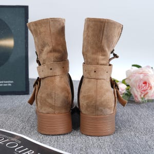 May include: A pair of brown suede ankle boots with a low block heel. The boots feature a buckle strap around the ankle and a zipper detail on the side. The boots are approximately 10 cm tall.