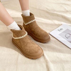 May include: A pair of brown suede ankle boots with a cream-colored crochet trim around the top. The boots have a thick, brown rubber sole. White socks are visible inside the boots. The word "Esty" is printed on a magazine in the background.