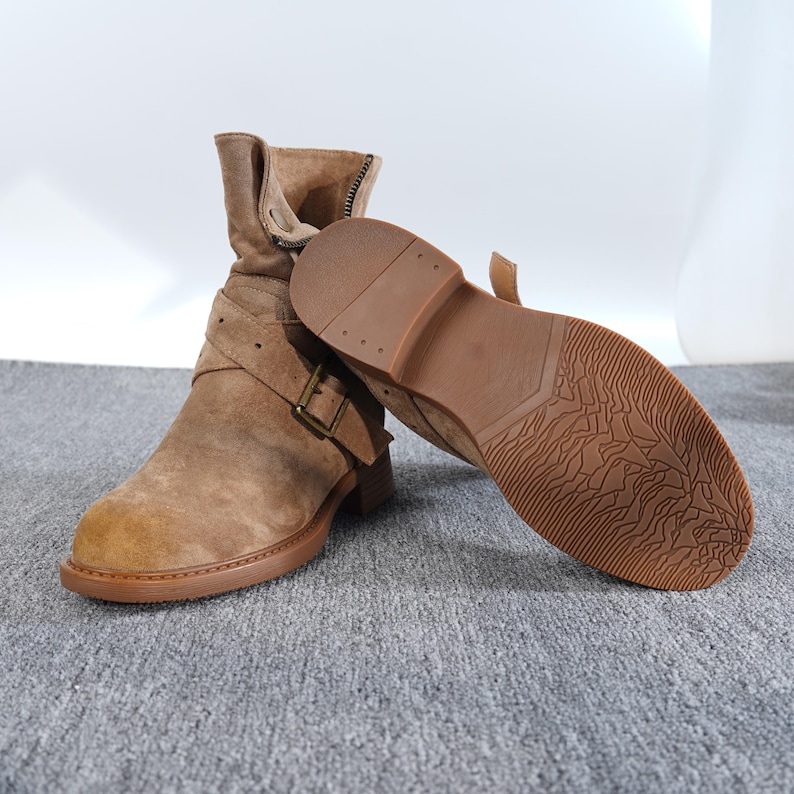 May include: Tan suede ankle boots with a low heel. One boot is upright, showing a buckle and strap detail, the other lies on its side, showcasing the textured sole. The boots have a zip and a rounded toe.
