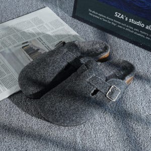 Pode incluir: Um par de tamancos cinzentos de feltro com fecho de fivela e sola de cortiça. Os tamancos são forrados com um material macio e de cor clara. O texto de fundo diz "SZA's studio al".