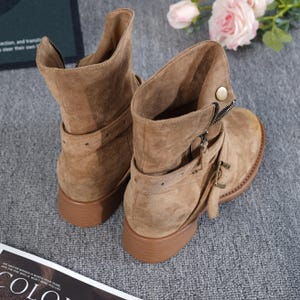 May include: Tan suede ankle boots with a low heel. The boots have a zip closure, buckle details, and a strap around the ankle. The boots are displayed on a grey surface.
