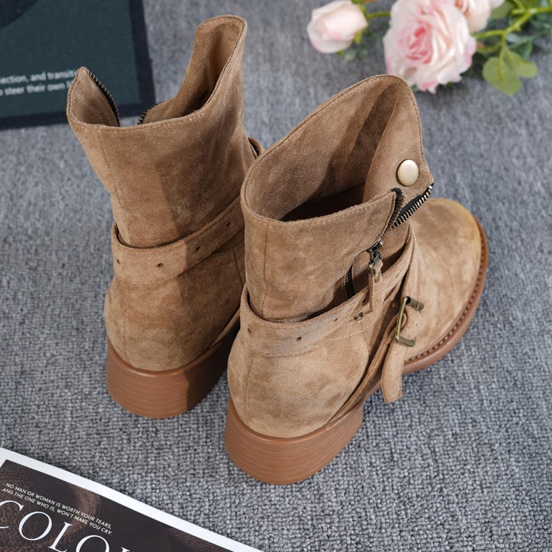 May include: Tan suede ankle boots with a low heel. The boots have a zip closure, buckle details, and a strap around the ankle. The boots are displayed on a grey surface.