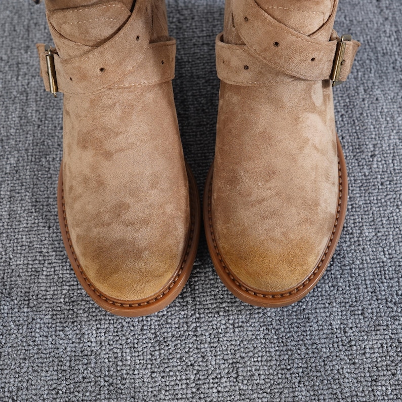 May include: A pair of tan suede ankle boots with rounded toes. The boots have a strap and buckle detail across the top of the foot and around the ankle. The soles are a darker brown colour.
