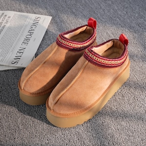 Puede incluir: Un par de zapatillas de ante color beige con una suela gruesa de color marrón claro. El cuello está adornado con una banda decorativa de bordados rojos y dorados. Hay un lazo rojo en la parte trasera de cada zapato. El texto "SINGAPORE NEWS" es visible.