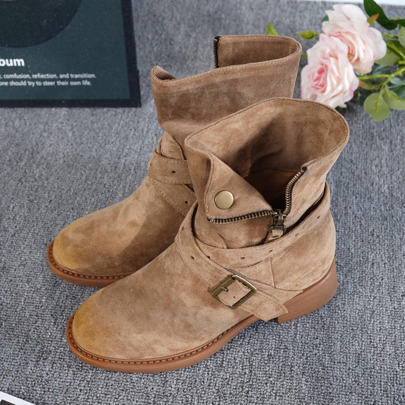May include: Tan suede ankle boots with a low heel. The boots have a side zip, buckle straps, and a round toe. The boots are displayed on a grey surface. The text in the image says: "bum, confusion, reflection, and transition, one should try to steer their own life."