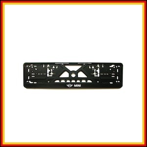 May include: A black rectangular license plate holder featuring a complex geometric cutout design and the white MINI logo centered at the bottom. The frame includes multiple mounting slots and holes for secure attachment to a vehicle exterior.