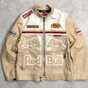 May include: Tan leather jacket with a cream-colored upper section and collar. The jacket features multiple patches, including "MOTUL", "MICHELIN", "BELL HELMETS", and "Esso". The front displays the "Red Bull" logo and text. A zipper and snap buttons secure the front.