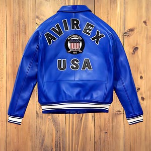 May include: A vibrant blue leather jacket with the word "AVIREX" arched across the shoulders, and "USA" below. The jacket features a patch with the words "ALL AMERICANS" and the year "1975". White stripes accent the cuffs and hem.