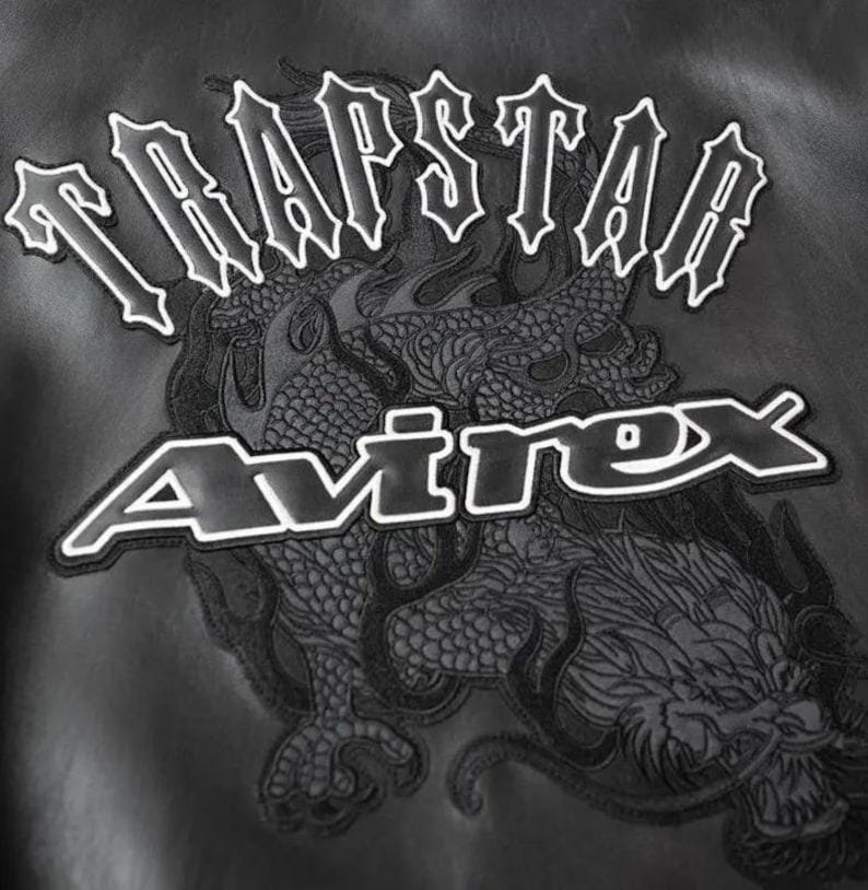 May include: Black leather jacket with white embroidered text that reads "TRAPSTAR" above "Avirex". A detailed dragon design is embroidered below the text, adding a bold, edgy aesthetic. The jacket's design is suitable for streetwear.