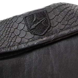 May include: Close-up of a black leather wallet. The top flap features a textured, scale-like pattern and a black embroidered emblem. The lower portion of the wallet is smooth black leather.