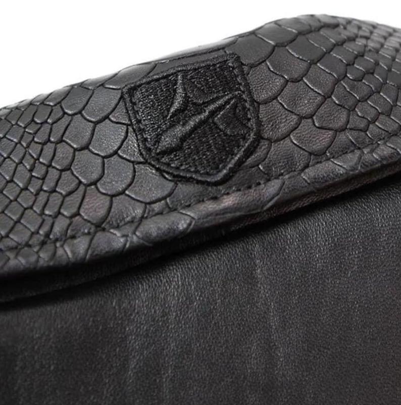 May include: Close-up of a black leather wallet. The top flap features a textured, scale-like pattern and a black embroidered emblem. The lower portion of the wallet is smooth black leather.