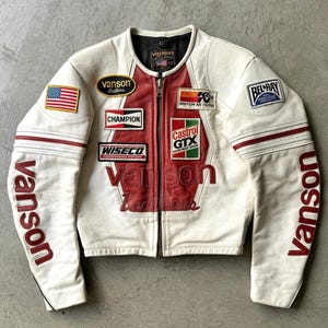 Może przedstawiać: Kremowa skórzana kurtka motocyklowa z czerwonymi akcentami i różnymi naszywkami. Kurtka posiada logo "Vanson" na rękawach i piersi, a także naszywki "Champion", "Castrol GTX" i flagę amerykańską. Styl vintage.
