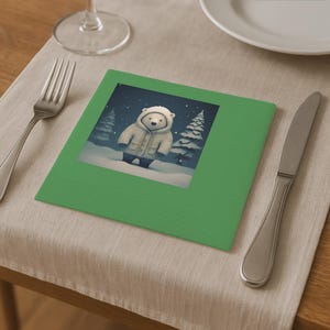 May include: A green paper napkin with a winter scene featuring a polar bear wearing a hooded coat. The bear stands in the snow with pine trees in the background. A fork, knife, and plate are on the table.