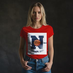 May include: Red t-shirt with a white square graphic featuring a navy blue letter "N" with a basketball inside. The graphic includes the text "EST. 1946" and "CHAMPS 2025".