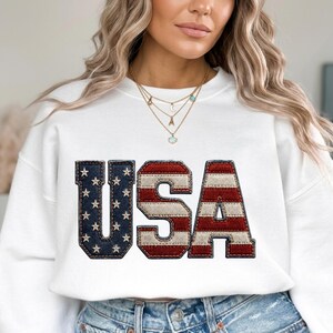 Könnte beinhalten: Weißes Sweatshirt mit dem Wort "USA" in einem Design im Denim-Stil. Die Buchstaben sind rot, weiß und blau, mit Sternen auf dem "U" und "S". Das Sweatshirt ist ein lässiger Stil.