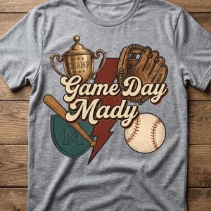 May include: Gray t-shirt featuring a baseball-themed graphic. The design includes a gold trophy, baseball glove, baseball bat, baseball, and the text "Game Day Mady". A red lightning bolt and a green patch with the letter "M" are also present.