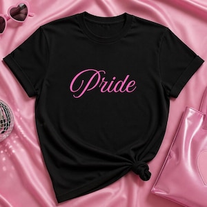 Puede incluir: Camiseta negra con la palabra "Pride" en escritura rosa. La camiseta está diseñada con un bolso rosa, gafas de sol en forma de corazón y una bola de discoteca sobre un fondo de satén rosa.