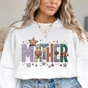 Puede incluir: Sudadera blanca con la palabra "MOTHER" en letras decorativas y coloridas. Las letras presentan estrellas, corazones y otros adornos. La sudadera es de cuello redondo.