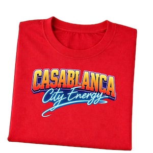 Op de afbeelding: Een opgevouwen rood t-shirt met de tekst "CASABLANCA City Energy" in een retro, gradiënt lettertype. De tekst is omlijnd in blauw en wit, met een schaduweffect. Het shirt is gemaakt van een zacht materiaal.