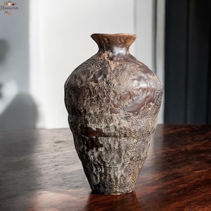 May include: A brown ceramic vase with a textured, uneven surface. The vase has a narrow neck and a bulbous body. The rustic vase is displayed on a dark wooden surface. The vase's height is approximately 20 cm.