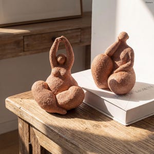Pode incluir: Duas esculturas de figuras sentadas em terracota. Uma figura tem os braços levantados acima da cabeça, a outra apoia um braço numa forma arredondada. As esculturas estão numa superfície de madeira e num livro. As figuras têm uma superfície texturizada.