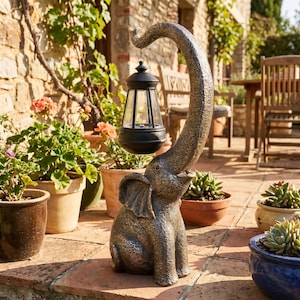 May include: A bronze-colored elephant statue with an extended trunk holding a black lantern. The elephant is seated, with its trunk curved upwards to suspend the lantern. The lantern has a classic design with clear panels. The statue is outdoors, surrounded by potted plants.