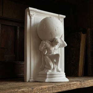 May include: A white Atlas statue bookend. The sculpture shows Atlas kneeling and supporting a globe. The bookend is set against a white architectural backdrop. The statue is on a wooden shelf with antique books in the background.