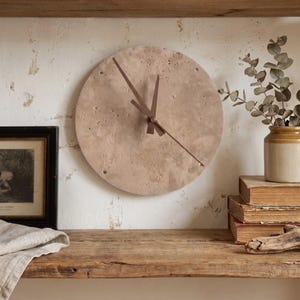 May include: A circular wall clock with a textured, light beige stone-like face and thin, dark brown metal hands. The clock features a porous, uneven surface finish and is mounted on a wall, displaying a simple design without numbers or markers.