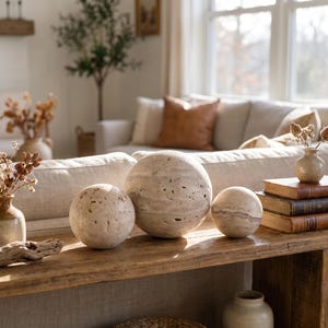 May include: Three spherical decorative objects made of beige, porous travertine stone with visible natural veining and pitted textures. The spheres are arranged in a row on a wooden surface, varying in size from small to large, showcasing organic patterns.