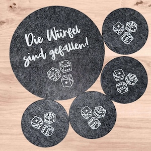 Könnte beinhalten: Ein Set aus fünf runden, dunkelgrauen Filzuntersetzern in verschiedenen Größen. Der größte Untersetzer zeigt den weißen Schriftzug Die Würfel sind gefallen! und drei Würfel. Die vier kleineren Untersetzer zeigen jeweils drei weiße Würfel.