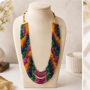 Op de afbeelding: Een meerstrengs ketting met kleine, gefacetteerde kralen in levendige tinten geel, groen, blauw en roze. De kralen zijn in een chevronpatroon over meerdere strengen gerangschikt en vastgezet met goudkleurige metalen eindkappen op een beige buste.