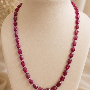 Puede incluir: Un collar con piedras preciosas ovales de color rojo intenso. Las cuentas están ensartadas, creando un bucle continuo. El collar se exhibe en un maniquí blanco, mostrando el color y el diseño de la joya. El fondo es de un tono suave y neutro.