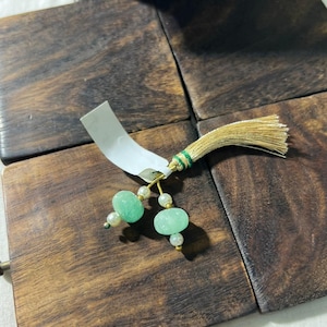 Puede incluir: Un adorno decorativo que presenta una borla de color dorado con un hilo verde, dos alambres dorados colgantes, cuatro cuentas blancas pequeñas y dos cuentas más grandes, redondeadas y de color verde claro translúcido, con una etiqueta.