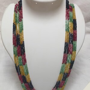 Puede incluir: Un collar de múltiples hilos con una variedad de cuentas de colores. El collar presenta hilos de cuentas rojas, verdes, amarillas y azules. El collar se exhibe en un busto blanco.