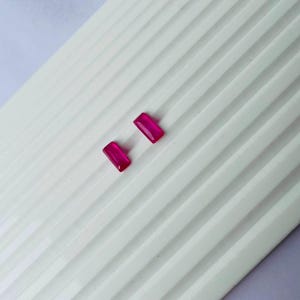 以下が含まれることがあります： 滑らかで光沢のある表面を持つ、鮮やかなフクシアピンク色の小さな長方形の宝石が2つ。これらのファセットカットされた石は透明感があり、平行な線が入った白い波状のテクスチャーを持つ表面の上に斜めに配置されています。