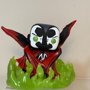 May include: A stylized vinyl figure of a character with a black and white mask, bright green eyes, and a red and black cape. The figure stands atop a translucent, light green base sculpted to resemble flickering flames or energy.