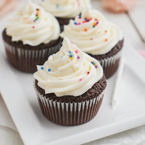 Könnte beinhalten: Vier Schokoladen-Cupcakes mit weißem Zuckerguss und bunten Streuseln auf einem weißen rechteckigen Teller. Die Cupcakes haben eine dunkelbraune Basis und befinden sich in weißen Papierförmchen. Der Zuckerguss ist gewirbelt und mit kleinen, bunten Streuseln bedeckt.