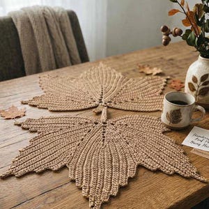Peut inclure: Deux sets de table en crochet beige en forme de grandes feuilles d'érable avec des coutures nervurées détaillées. Une tasse en céramique avec un motif de feuille, un vase en céramique avec des branches séchées et une petite carte rectangulaire avec du texte manuscrit sont également présents.