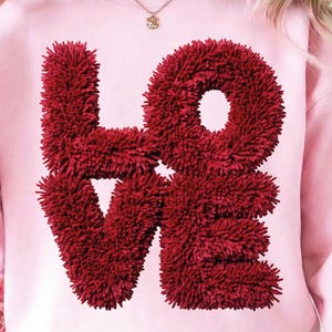 May include: A light pink sweatshirt with the word "LOVE" in a textured, deep red font. The letters appear to be made of a fuzzy material, creating a 3D effect. The sweatshirt is a casual style, perfect for Valentine's Day or everyday wear.
